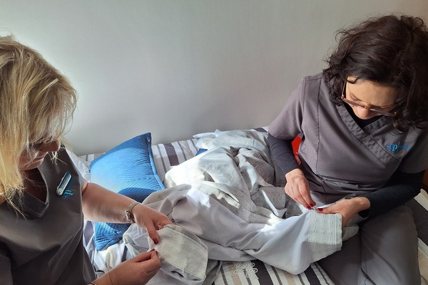 Two women in uniforms are sitting on a bed, carefully folding or inspecting light-colored clothing or linens in a neatly made room with striped bedding and a blue pillow.