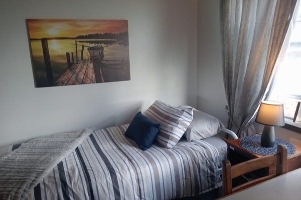 A neatly made single bed with striped bedding and several pillows is positioned next to a wooden nightstand with a lamp and a window with sheer curtains; a framed sunset photograph hangs on the wall above the bed.