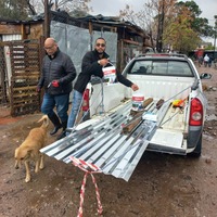 Two men are unloading sheets of corrugated metal from the back of a white bakkie in a rustic outdoor area, with a tan dog standing nearby and informal structures in the background.