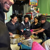 everal adults are serving food from a large pot to a child seated at a table inside a cozy, colorful room, while others look on and assist