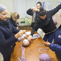 Five people are gathered around a table, working with large balls of wool and knitting needles, appearing to be engaged in a group knitting or crafting activity.