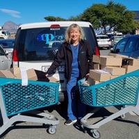 A smiling woman stands between two shopping carts filled with cardboard boxes in a parking lot, with a white van behind her displaying a blue logo on the rear window.