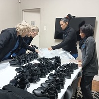 Four people are standing around a table covered with rows of black shoes, appearing to sort or organize them in a room with a television on the wall.