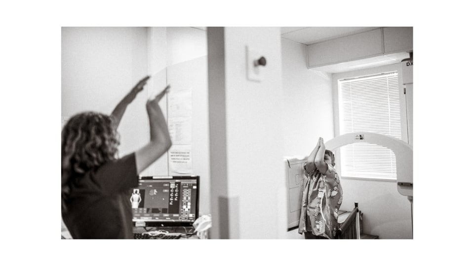 A radiographer stands beside a computer monitor, raising their arms to demonstrate a pose to a young patient, who is mimicking the pose under an X-ray machine in a medical imaging room. (Radiographers are healthcare professionals trained to perform diagnostic imaging examinations such as X-rays, CT scans, and MR scans.)