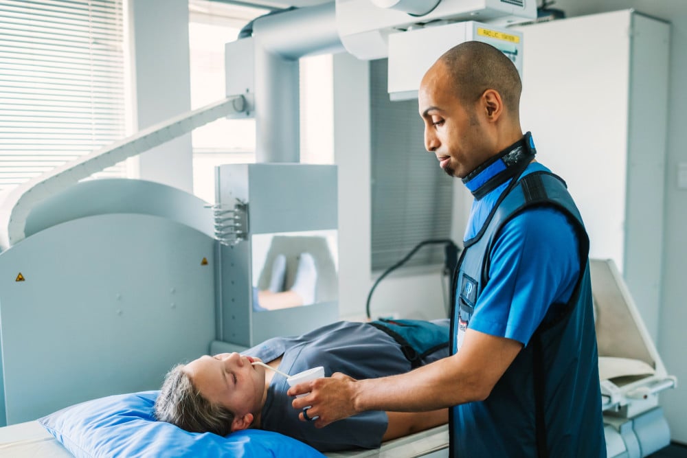 A radiographer in a protective vest prepares a patient lying on a table under a medical imaging device, likely an X-ray or CT scanner, in a bright clinical setting.