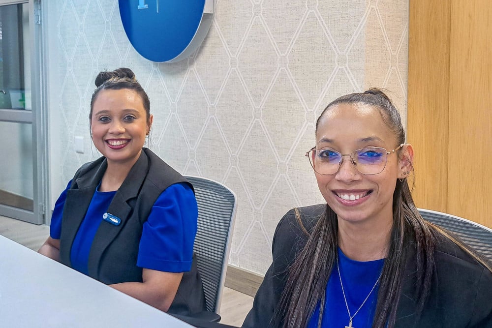 Two smiling medical receptionists, Nikita and Marilu Roberts, seated at the front desk of SCP Radiology Panorama, wearing blue uniforms and black vests.